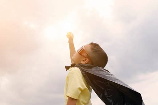 boy in superhero cape with arm to sun and sky